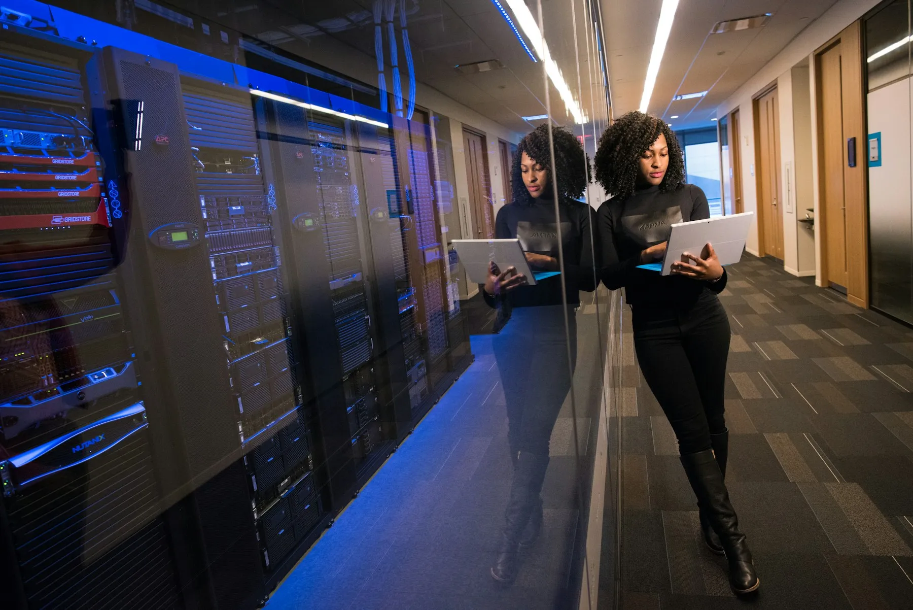 An engineer with a laptop walking past server racks lit in blue.